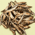 A pile of dried small fish, arranged on a solid pale yellow-green background.
